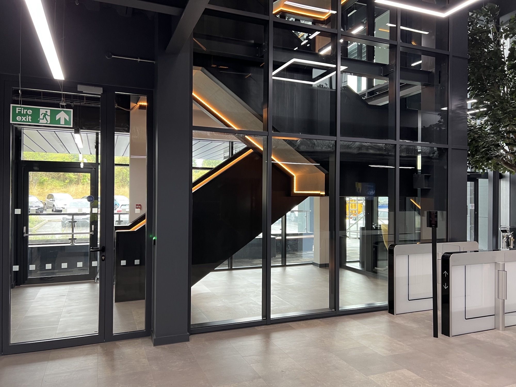 Modernes Gebäude mit Glaswänden, schwarzer Treppe mit LED-Beleuchtung und einem Eingang mit Drehkreuz